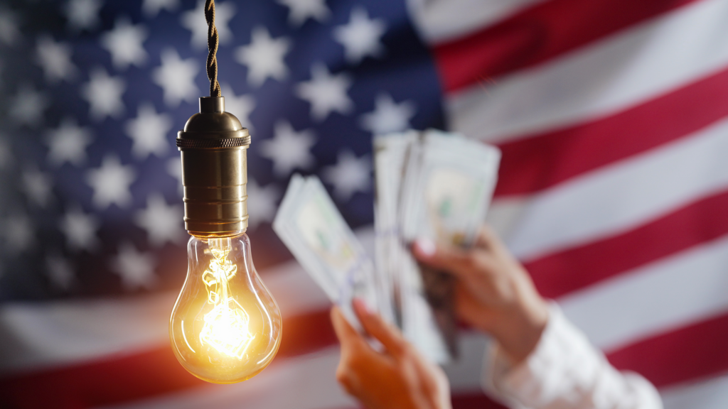 hands shown holding money with a light bulb in the foreground and an American flag in the background