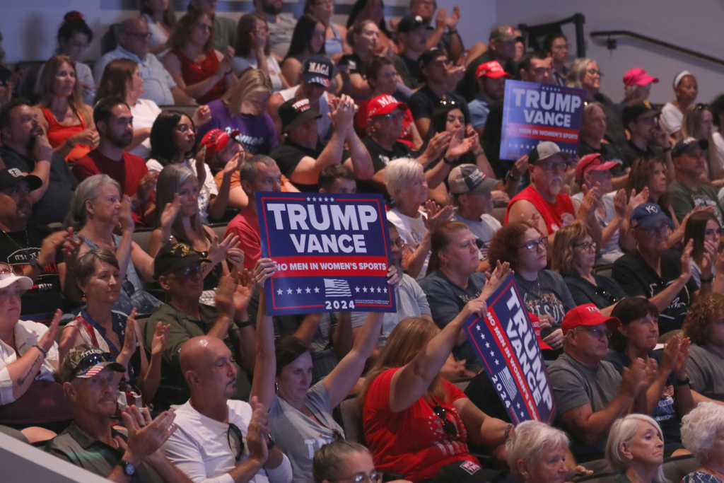 A crowd of people attending a political event. Some people are holding up Trump campaign signs.